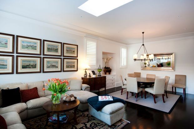 Bright, refined open-plan living and dining room with white walls, decorative crown moulding, dark timber flooring, and a large skylight. A generous sectional sofa, upholstered ottoman, round timber coffee table, framed artwork gallery, sideboard, dining table, upholstered dining chairs, and a traditional chandelier create a comfortable traditional interior. A wall mirror reflects part of the room, while plantation shutters provide window privacy.
