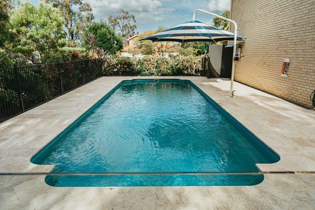 A rectangular in-ground swimming pool occupies a paved outdoor courtyard or backyard. The pool has turquoise water, pale stone or concrete surrounds, a dark metal safety fence, a large striped cantilever-style umbrella, and a light tan brick exterior wall. Mature landscaping and trees are visible beyond the fence.