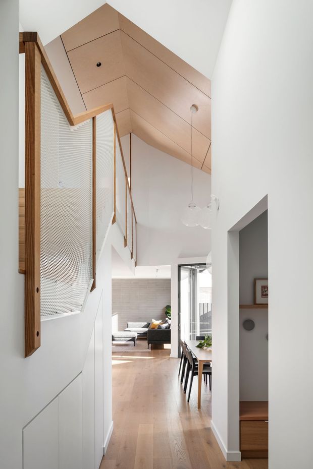 A bright, narrow residential hallway or circulation space with pale engineered timber flooring, white walls, and a sculptural warm timber-lined ceiling that follows the roof form. On the left is a tall timber-framed perforated screen or balustrade, while the right opens toward a compact stair or built-in area. The hallway leads into an open-plan dining and living space with a timber dining table, black chairs, sofa, and glazing. Minimal contemporary detailing, abundant natural light, and integrated timber joinery create a warm, refined interior.