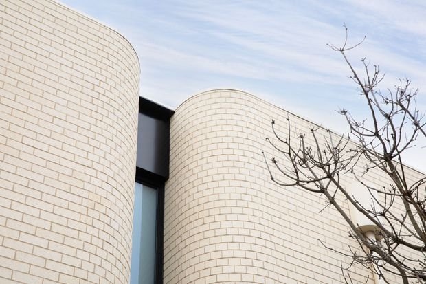 Exterior architectural detail showing curved, cylindrical walls clad in pale cream brickwork, with dark-framed glazing recessed between the rounded forms. A leafless tree overlaps the facade, and the building has a sculptural contemporary composition beneath a bright sky.