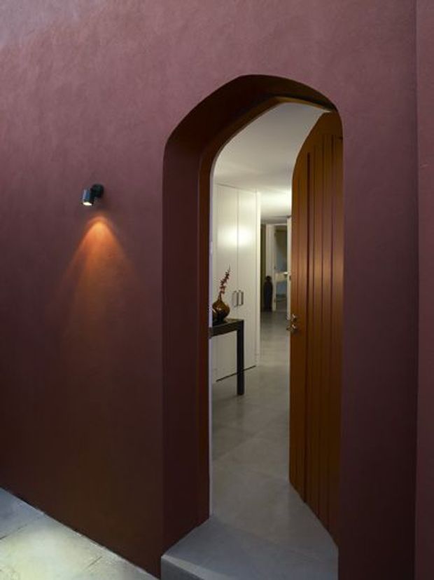 Interior view of a sculptural arched doorway with a tall fluted timber door opening into a bright hallway. The surrounding walls are finished in a deep muted mauve or burgundy tone, with a small directional wall light casting a warm glow. A grey tiled or stone floor continues through the threshold, and a narrow console table with decorative objects is visible beyond.