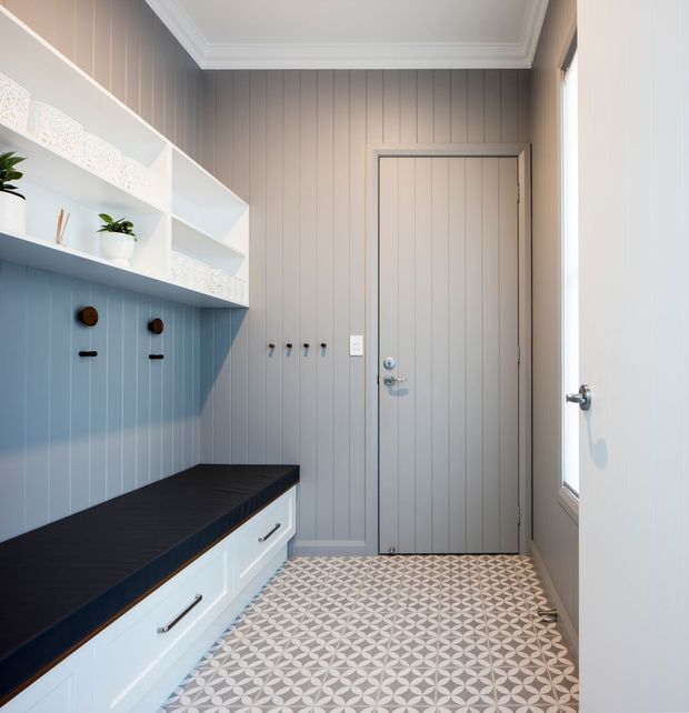 Bright, compact entryway with grey vertical wall panelling, a built-in white storage bench and drawers topped with a dark benchtop, open display shelving, coat hooks, and a patterned geometric tiled floor. A pale grey panelled door and side glazing bring in natural light. The space has a clean, contemporary coastal feel with traditional cornice detailing.