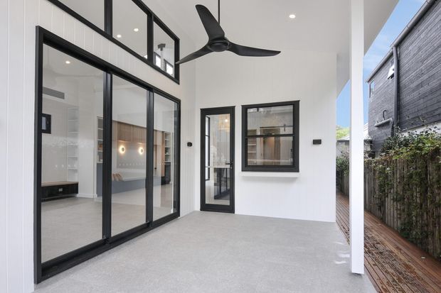 A bright, contemporary covered outdoor living area or terrace with a smooth light-grey floor, white walls, black-framed floor-to-ceiling sliding glass doors, and a matching black-framed window and hinged door. A sculptural black ceiling fan hangs overhead. The glazing opens toward a visible minimalist interior, while a narrow timber deck and neighbouring dark-clad structure are visible to the side.