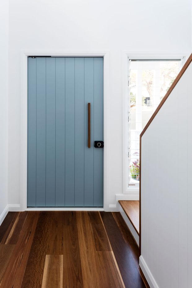 Bright, minimalist residential entryway featuring a tall blue-grey vertically panelled front door with a slim timber handle and black electronic lock. The entry has dark timber flooring, white walls and trim, a narrow window beside the door, and a staircase with white balustrade panels and timber edging.