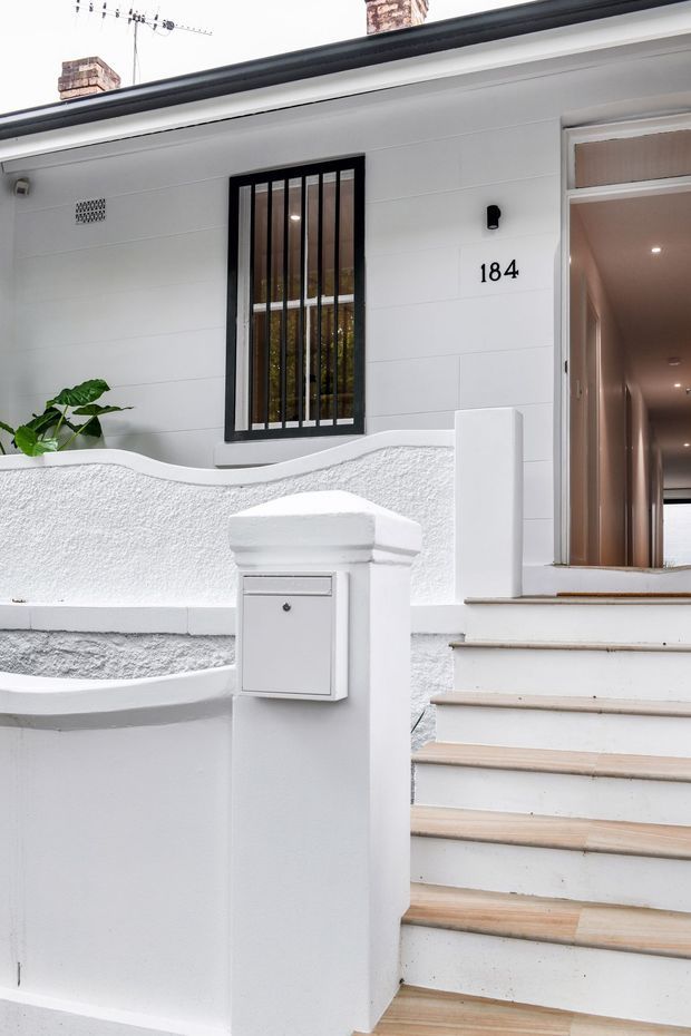 Exterior view of a compact residential entryway with white rendered walls, a curved textured boundary wall, front steps, black barred window, open front door, and a white letterbox pillar. The house number 184 is visible beside the entrance, with a hallway glimpsed inside.