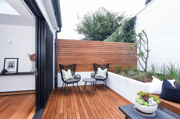 A compact, modern outdoor deck courtyard viewed from inside through fully opened black-framed glazing. The deck has warm timber boards, two black wire-frame lounge chairs with white cushions, a small round table, built-in bench seating with cushions, and a raised white planter bed against a horizontal timber privacy screen. A portion of the adjacent minimalist interior is visible.
