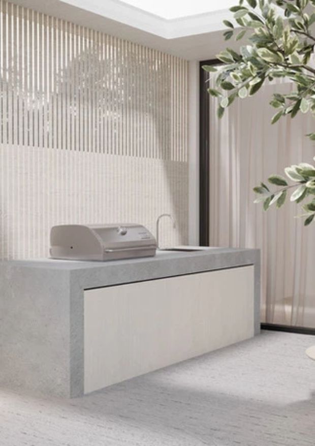A refined, minimalist outdoor kitchen with a compact built-in barbecue set into a pale stone island. The space features light vertical screening, pale timber or wood-look cabinetry, a light textured floor, and expansive glazed openings with a leafy branch in the foreground. The overall palette is soft and neutral with clean contemporary detailing.