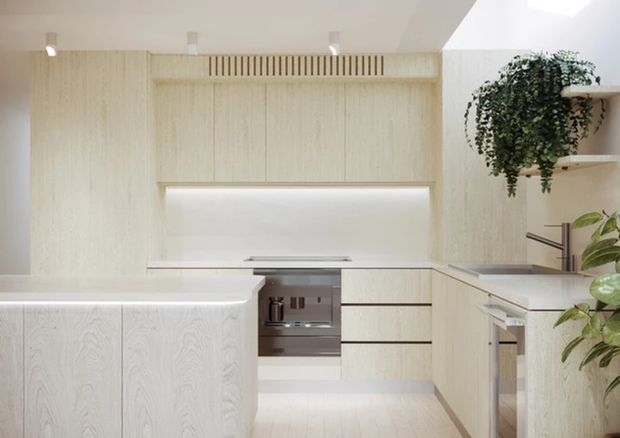 A bright, compact contemporary kitchen with pale timber-grain cabinetry, white benchtops and splashback, integrated stainless-steel appliances, and a peninsula island. Under-cabinet lighting, vertical ventilation detailing, open shelving, and trailing indoor plants add subtle visual interest.