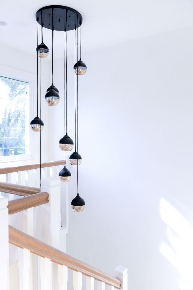 Bright, minimalist stairwell with white walls and balustrades, warm timber handrails, and a dramatic multi-light pendant installation hanging from a round black ceiling plate over the stair void. Natural light enters through a tall window, creating a clean, airy atmosphere.