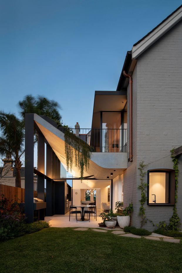 A dusk view of a contemporary residential courtyard and rear facade. The two-storey home combines a white-painted brick wall with dark metal-framed glazing, projecting balconies, and a sculptural covered outdoor dining area. Warm architectural lighting illuminates the terrace, kitchen-like outdoor counter, planting, and lawn, while a curved concrete balcony edge and slim black balustrades add modern detailing.