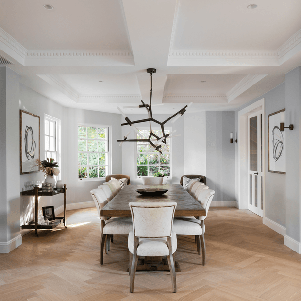 Bright, refined dining room with a long rectangular timber dining table surrounded by upholstered dining chairs. The room features pale blue-grey walls, white decorative coffered ceilings with ornate cornice detailing, herringbone timber flooring, tall multi-pane windows, framed abstract artwork, and a sculptural black linear pendant light.