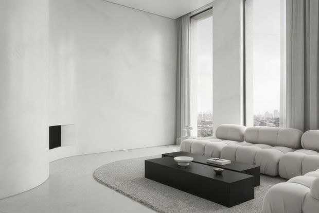 A minimalist, monochromatic living room with curved white walls, floor-to-ceiling windows, grey curtains, and expansive city views. A large modular white sectional sofa surrounds a low black rectangular coffee table, set on a pale textured rug over a light grey floor. The room is bright, spacious, and refined, with sculptural rounded forms and very little decoration.