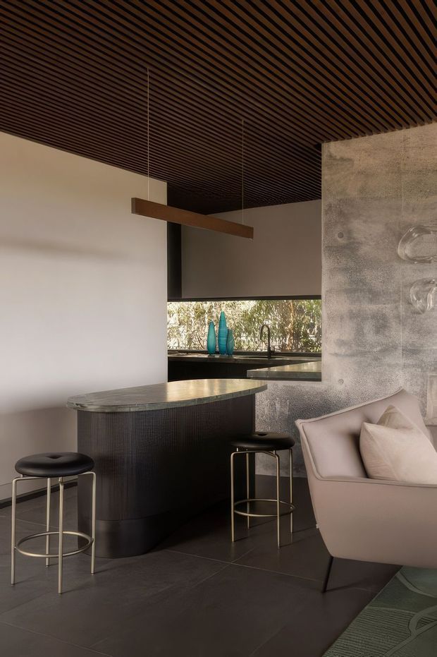 Sophisticated contemporary interior showing a compact curved bar or kitchen island with dark ribbed cabinetry and a stone benchtop, paired with black and brass stools. A dark timber slatted ceiling, polished concrete-textured feature wall, concealed cabinetry, pendant light, and pale upholstered seating create a refined, moody lounge-bar atmosphere. A window provides greenery beyond the room.