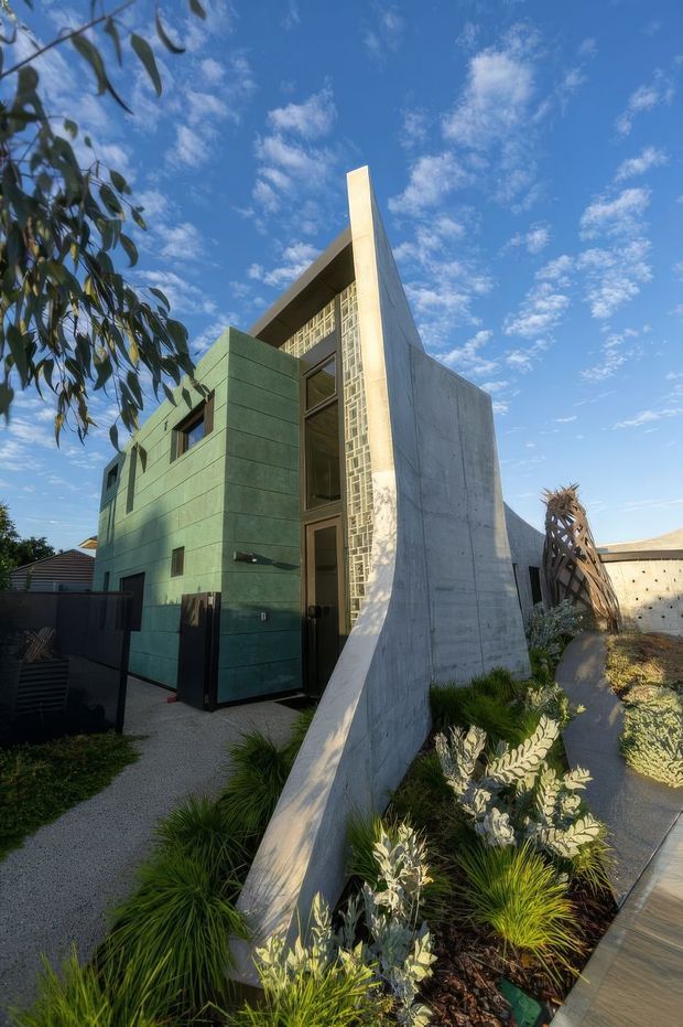 Exterior view of a distinctive contemporary concrete residence with a tall angular sculptural wall, green horizontal-panel cladding, narrow vertical glazing, and patterned glass blocks. The house is set within a landscaped front garden with gravel paths and drought-tolerant planting under a bright blue sky.