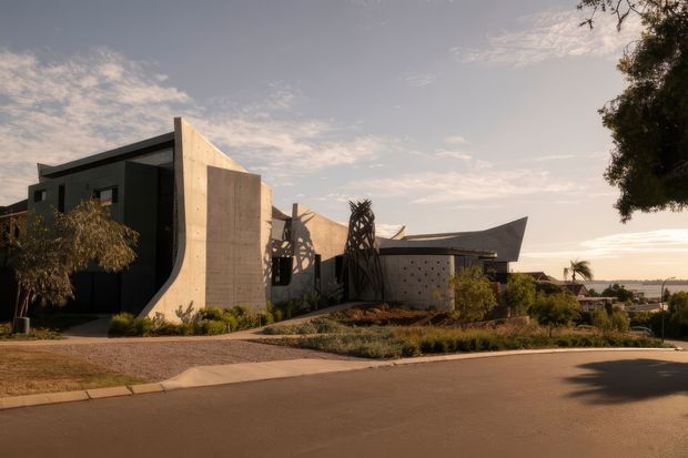 Street-level exterior view of a sculptural contemporary residence composed of dark geometric volumes, exposed concrete walls, angled roof forms and perforated screening. The landscaped frontage includes low planting and paved pedestrian access, with a broad water or coastal horizon visible beyond the property.