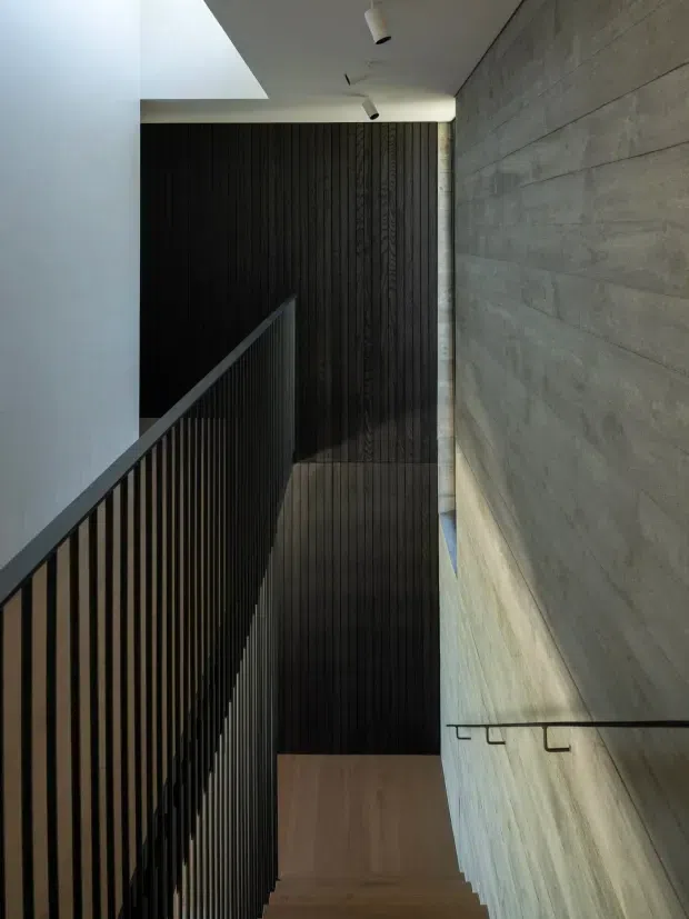 A contemporary interior stairway viewed looking downward, with pale timber stair treads, a slim black metal balustrade, a dark vertically grooved timber feature wall, and a textured grey concrete or masonry wall. Recessed and surface-mounted spotlights illuminate the tall, narrow stairwell, creating a restrained, dramatic atmosphere.