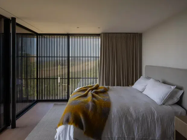 A contemporary bedroom with a large upholstered bed, white bedding, and a mustard-yellow throw. Floor-to-ceiling glazing with dark vertical screening frames the left side, while full-height taupe curtains cover the rear wall. The room has a restrained neutral palette, recessed ceiling lighting, and a soft area rug.