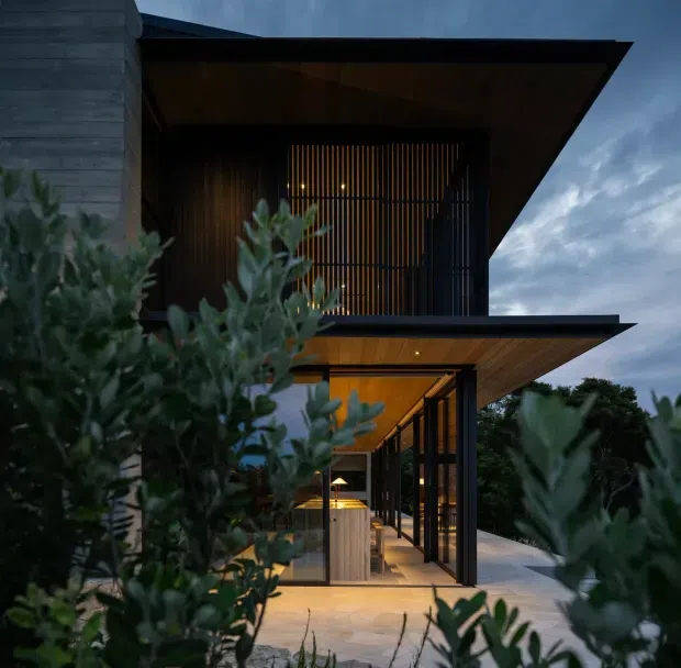 Twilight exterior view of a contemporary multi-level residence with dramatic cantilevered flat roof planes, dark vertical screening, warm timber soffits, extensive glazing, and a partially visible illuminated outdoor living or entry area. Dense foreground planting frames the building.
