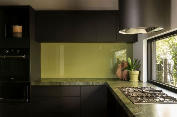 A compact, dark-toned contemporary kitchen with matte black floor-to-ceiling cabinetry, integrated appliances, a green glass splashback, and a green-veined stone benchtop. A gas cooktop sits beneath a sculptural stainless-steel rangehood beside a large black-framed window, with potted plants adding natural detail.