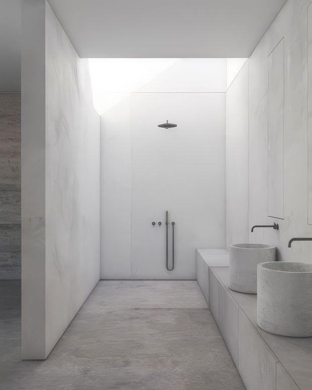 Minimalist monochromatic bathroom with a walk-in shower, wall-mounted shower fittings, dual cylindrical vessel basins, and a long integrated vanity. Large pale stone-look wall panels and flooring create a seamless, sculptural interior, with soft natural light entering from an overhead or clerestory opening.