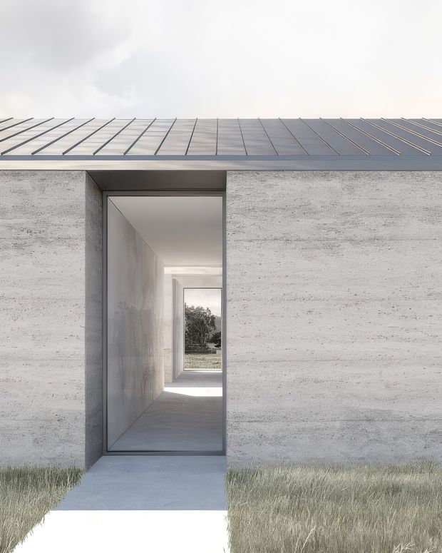 Minimalist single-storey concrete building exterior with a flat metal roof and a deeply recessed, portal-like entrance. The rough pale concrete facade contrasts with the dark roof edge and narrow glazed passage leading through the building. Long grasses and a simple concrete path frame the entrance, creating a stark, geometric composition.