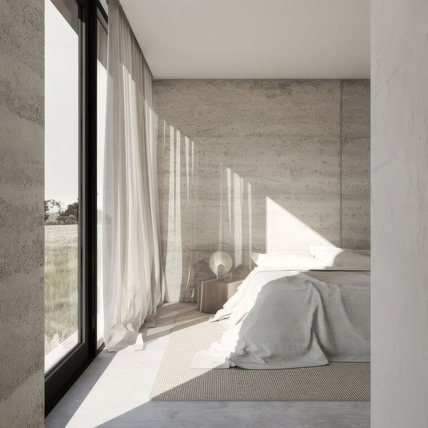 A serene minimalist bedroom with a low bed dressed in white linen, set against raw textured concrete walls. Full-height black-framed glazing with sheer curtains runs along the left side, casting strong geometric sunlight and shadows across the pale floor and woven rug. A small timber bedside table and sculptural lamp sit beside the bed, with a grassy landscape visible outside.