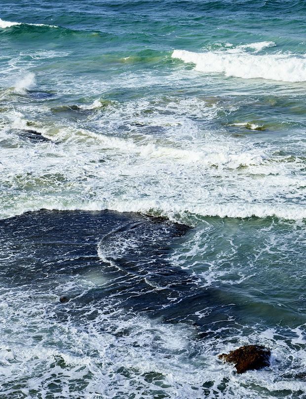 A close-up view of waves breaking over dark rocky coastal outcrops, with swirling white sea foam and blue-green ocean water. No building or architectural subject is visible.