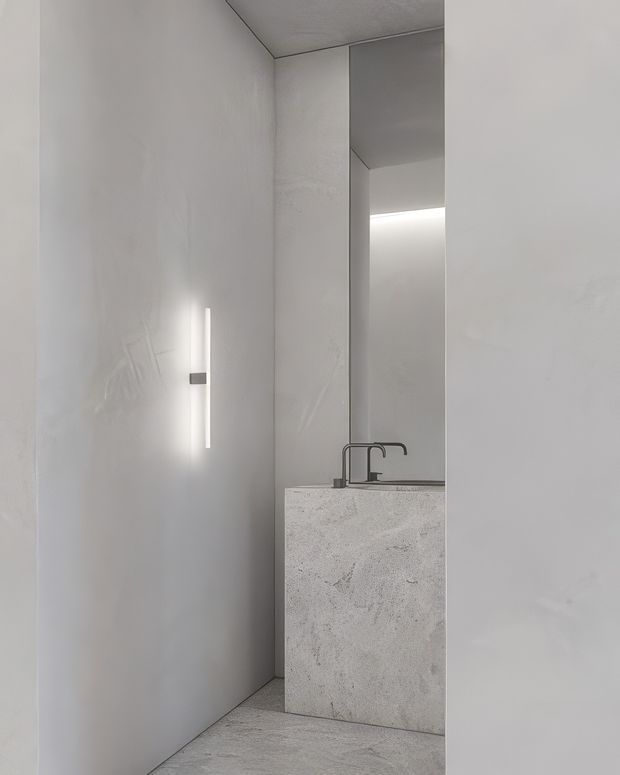 A minimalist bathroom interior in pale grey and white tones, featuring a monolithic stone vanity with an integrated basin and dark matte tapware. Full-height wall and floor surfaces create a seamless, sculptural appearance. A tall mirror and concealed linear lighting add depth and a soft glow, while a slim vertical wall light provides a bright architectural accent.