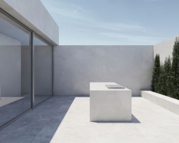Minimalist rooftop or courtyard terrace with pale rendered walls, large frameless-looking glazed sliding doors, light stone or concrete paving, and a simple white built-in outdoor kitchen island with a cooktop. Tall narrow evergreen planting lines the edge, creating a quiet, highly restrained architectural setting under a bright sky.