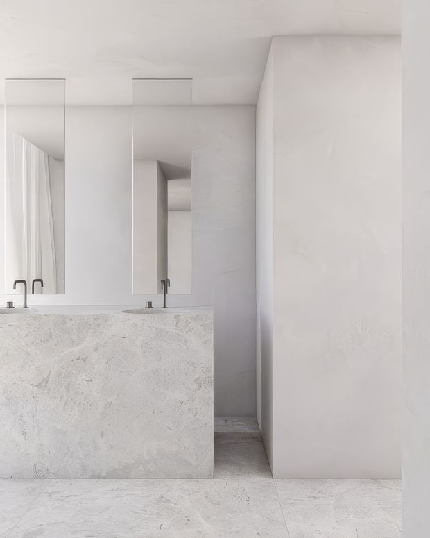 Minimalist monochromatic bathroom featuring a large freestanding or built-in concrete-look vanity with integrated basins and slim dark tapware. Tall frameless mirrors reflect the vanity and sheer curtains, while pale textured wall surfaces and large light stone-look floor tiles create a seamless, sculptural interior.
