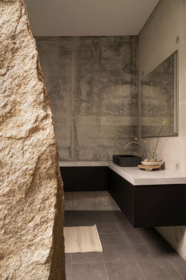 A compact contemporary bathroom with a dramatic rough natural-stone partition wall, large grey concrete-look wall and floor tiles, a wall-mounted black vanity with a pale stone benchtop, integrated basin, curved brass tapware, and a large rectangular mirror. The space has warm, earthy tones and a sculptural, minimalist fit-out.