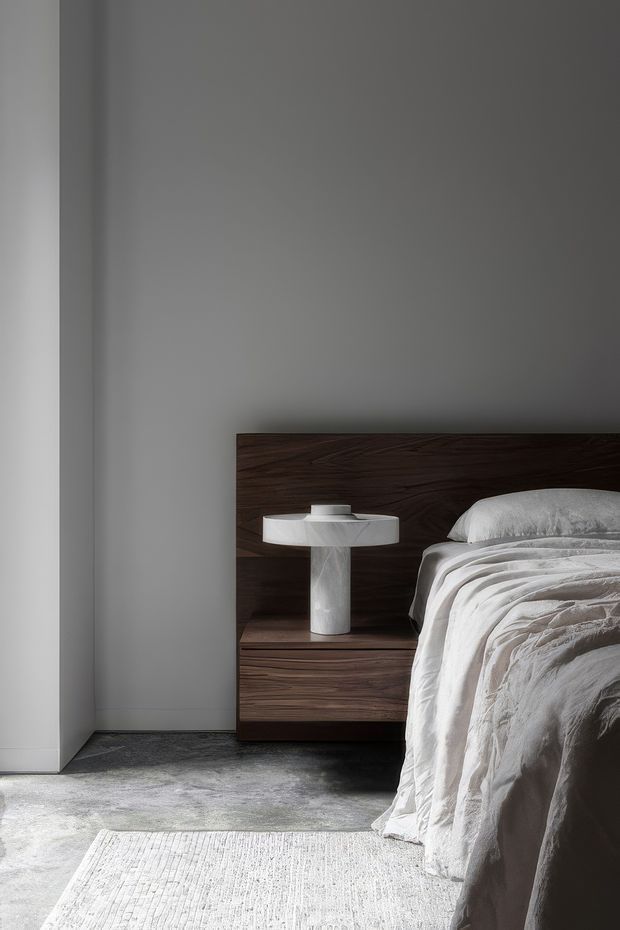 A softly lit minimalist bedroom vignette featuring a dark wood bedhead integrated with a floating bedside cabinet, a sculptural white table lamp, and white textured bedding. The room has smooth pale grey walls, a dark grey floor, and a light textured rug, creating a restrained, contemporary atmosphere.