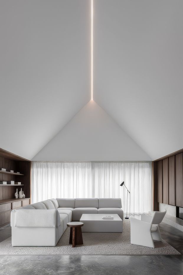 A refined contemporary living room with a dramatic vaulted ceiling and a slim linear recessed light at the ridge. Floor-to-ceiling sheer curtains span the rear wall, while dark timber built-in cabinetry frames both sides. The room contains a large pale sectional sofa, a low light coffee table, sculptural white lounge and dining chairs, a slim floor lamp, and a textured neutral rug over a dark polished floor. The overall composition is minimalist, calm, and monochromatic with warm timber accents.