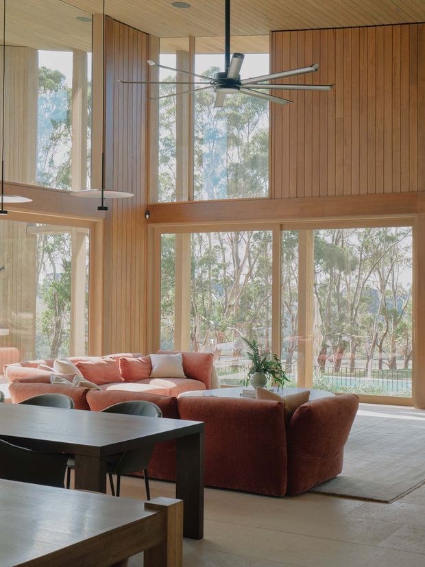 A spacious, warm-toned open-plan living and dining area with dramatic floor-to-ceiling glazing overlooking trees. The room features extensive vertical timber wall panelling and a matching timber ceiling, exposed structural columns, a large contemporary ceiling fan, sculptural pendant lights, a coral-toned sectional sofa, rounded lounge chairs, a dark dining table, and a pale tiled or stone floor. The high glazing and timber-lined volume create an airy, nature-connected interior.