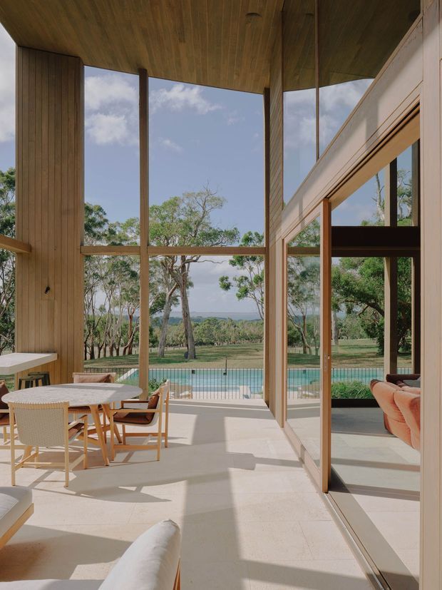 A bright, double-height glazed living and dining space opening toward a swimming pool and landscaped rural setting. The room features extensive floor-to-ceiling glazing with slim timber frames, vertical timber wall panelling, a timber-lined ceiling, pale stone or concrete flooring, a dining table with woven chairs, and a warm-toned sofa visible in reflection. The design is airy and strongly connected to the outdoors.