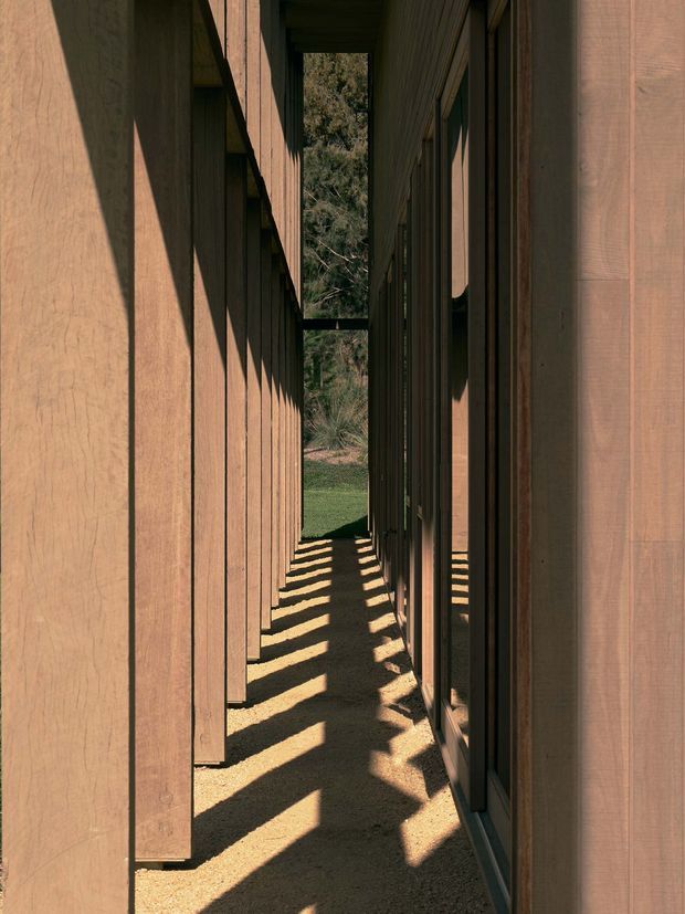 A narrow exterior passageway between two parallel timber-clad building volumes. Vertical warm-toned wood boards line both sides, with dark-framed glazing and deep recesses along the right-hand wall. Strong sunlight creates repeated diagonal shadows across the compact gravel or earthen walkway, which opens toward a small lawn and dense trees at the far end. The composition emphasizes a sheltered, linear architectural connection and rhythmic facade detailing.