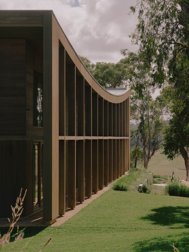 Exterior view of a contemporary timber-clad building in a rural, landscaped setting. The long facade features closely spaced vertical timber fins or posts, recessed glazing, and a distinctive gently curving roofline or canopy that creates a sheltered verandah-like edge. Mature trees, lawn, and open countryside surround the structure.
