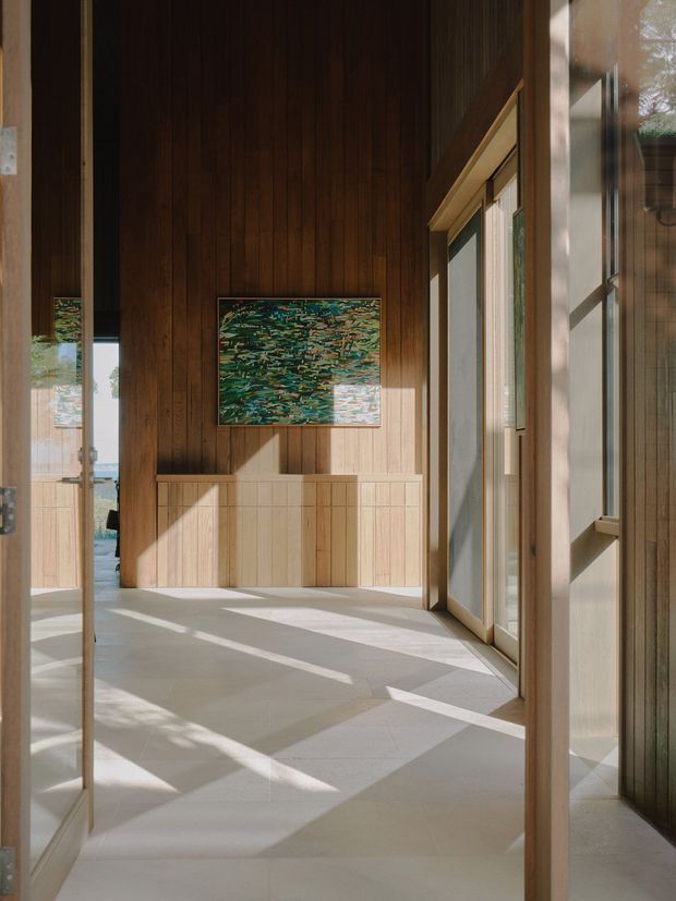A sunlit interior hallway or entry space with warm vertical timber wall panelling, a built-in timber console or storage unit, pale large-format tiled flooring, and extensive glazed doors or windows along the right side. Strong angular sunlight creates geometric shadows across the floor and walls. A colourful abstract artwork provides the main visual accent, while glimpses of another room and exterior landscape are visible through openings and glazing.