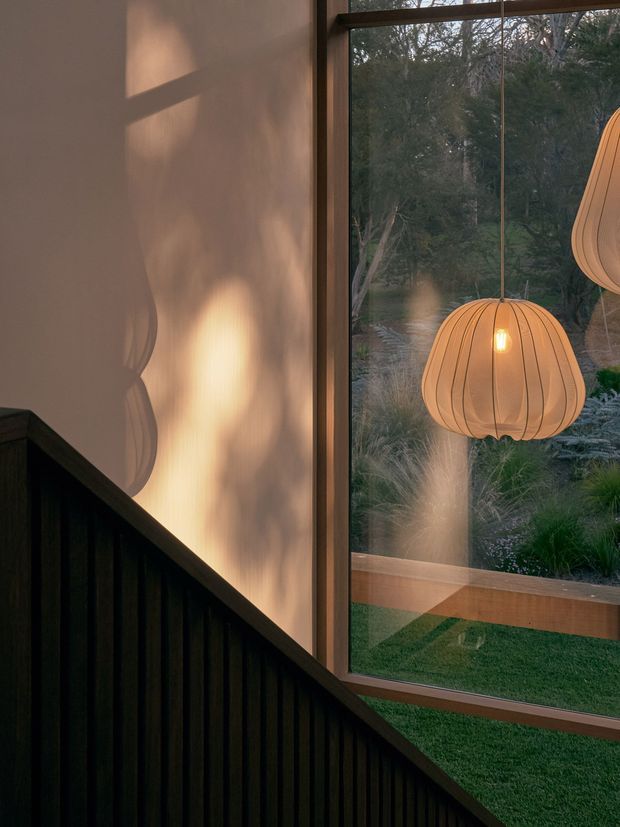 Moody architectural interior detail showing a dark timber-clad stair or wall rising diagonally beside a large floor-to-ceiling window. Warm sculptural pendant lights hang in front of the glazing, with their forms reflected on the plaster wall and window. The view beyond shows a landscaped garden and trees, while timber window framing and warm evening light create a dramatic, intimate atmosphere.