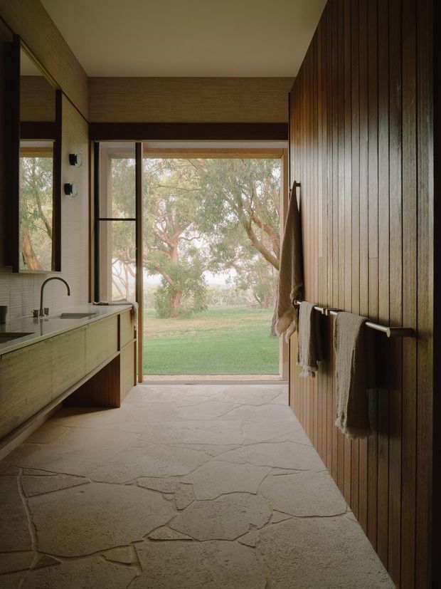 A warm, narrow bathroom with a long floating vanity and integrated basin on the left, large mirror, and a full-height glazed door opening directly onto a garden. The right wall is clad in vertical dark timber battens with simple towel rails and hanging towels. The floor has irregular large-format natural stone slabs, while the ceiling and upper walls are restrained and neutral. The room has a calm, earthy, indoor-outdoor character with abundant natural light.