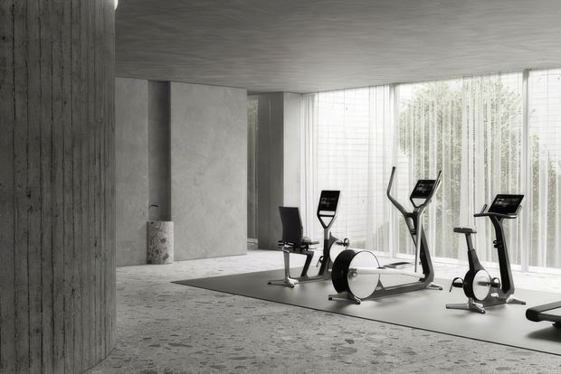 A minimalist, monochromatic home gym with stationary exercise bikes and cardio equipment arranged on a dark exercise mat. The space has exposed grey concrete walls, columns, ceiling, and textured concrete flooring, alongside expansive floor-to-ceiling glazing with sheer curtains and filtered greenery beyond. The restrained palette and abundant diffuse daylight create a refined, calm atmosphere.