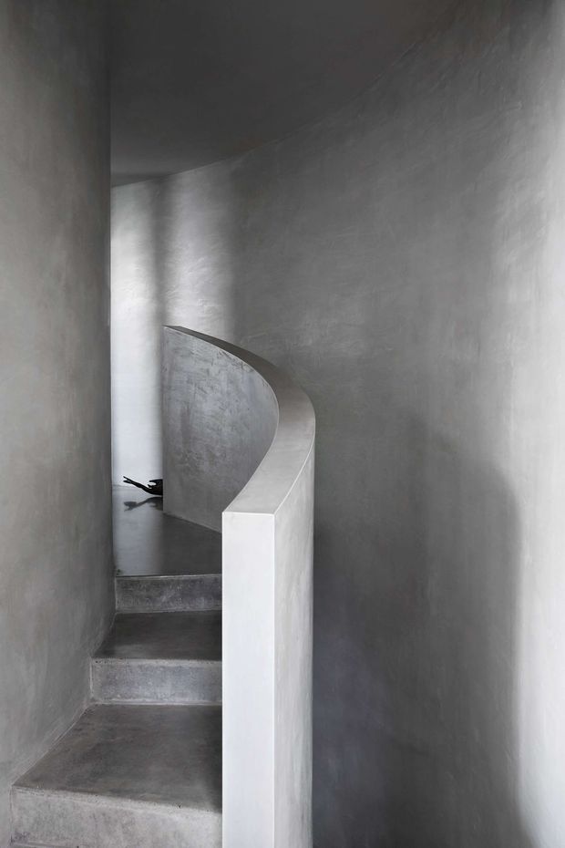 A narrow, sculptural interior stairway with raw grey concrete steps and continuous curved parapet. The walls and ceiling are finished in a seamless mottled grey material, creating a monochromatic, minimalist architectural composition with soft natural light.