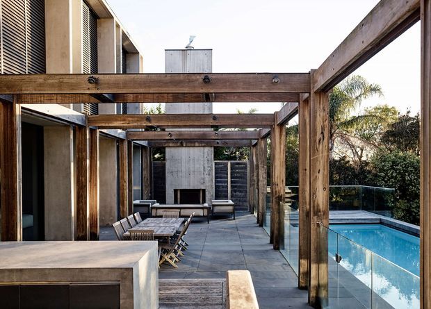 A contemporary outdoor entertaining terrace beside a swimming pool, framed by a substantial timber pergola with exposed beams and posts. The paved terrace includes an outdoor dining setting, lounge seating, a built-in concrete outdoor kitchen or barbecue bench in the foreground, and a glass pool barrier. Concrete and timber architectural finishes create a warm, refined indoor-outdoor connection.