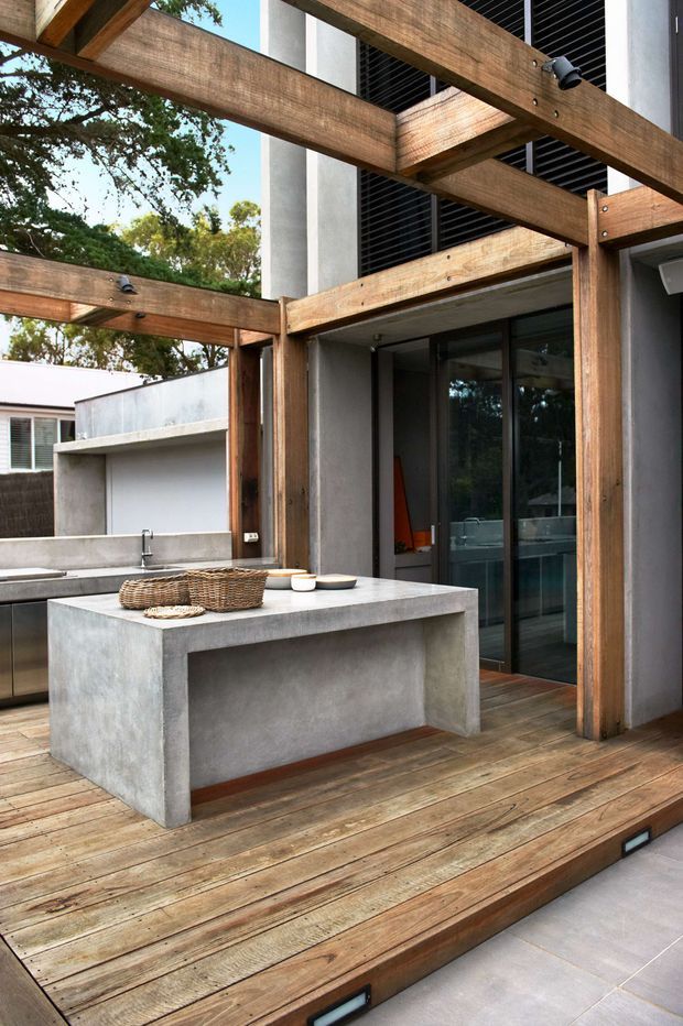 A contemporary outdoor kitchen and entertaining area on a timber deck. The space features a substantial concrete island bench, matching concrete outdoor counter and sink, exposed timber pergola beams with integrated spotlights, dark-framed sliding glass doors, and grey rendered walls. Timber decking steps down to a pale tiled surface, with greenery visible beyond.