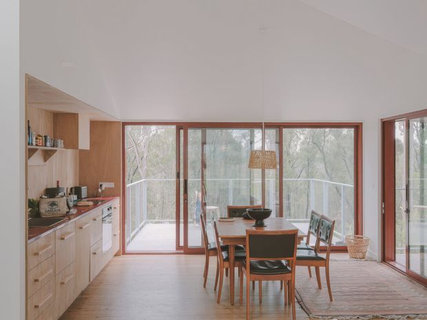 Bright open-plan kitchen and dining area with extensive warm timber cabinetry, integrated shelving, a timber dining table and mid-century-style chairs. Full-height timber-framed glazing opens onto a deck surrounded by trees, while a woven pendant light and neutral finishes create a warm, airy atmosphere.