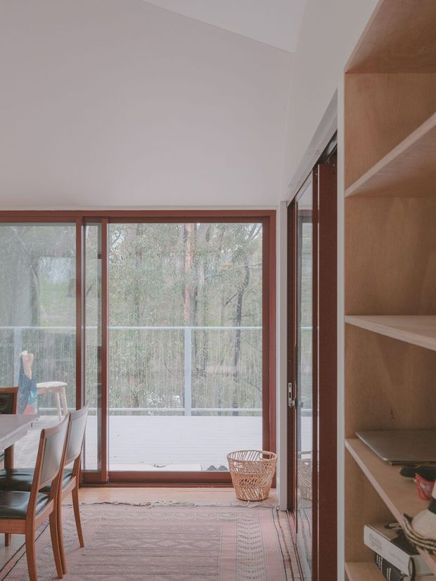 A bright, quiet interior corner with a timber-framed glazed door opening onto a balcony and wooded landscape. A dining table and chairs are partially visible on the left, while open light-wood shelving occupies the right side. The room has white walls and ceiling, warm timber framing, a patterned rug, and soft natural light.