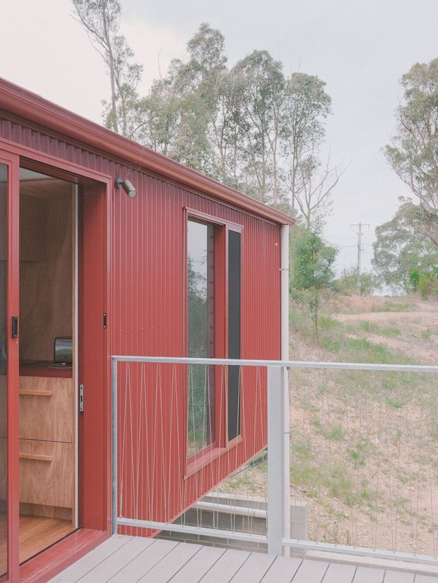 Exterior view of a compact red corrugated-metal-clad building with a small raised deck and wire-panel railing. A sliding glass door opens to a warm timber-lined interior, while tall eucalyptus trees and a dry rural landscape surround the structure.