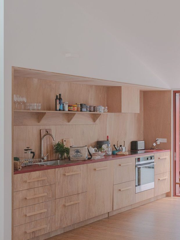 A compact, contemporary kitchen with a full-height light timber veneer cabinetry wall, integrated open shelving, and long pale timber lower cabinets. The kitchen includes a dark red-toned benchtop, stainless-steel sink and tap, cooktop, built-in oven, small appliances, and glassware. Warm timber flooring and a red-framed glazed door or window appear at the right edge. The space is softly lit and has a warm, understated feel.