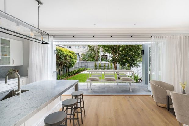 Bright open-plan kitchen and dining area opening through fully glazed sliding doors to a landscaped backyard. The room features a pale stone island with seating, white cabinetry, timber flooring, a contemporary linear pendant, and a dining table positioned beside the garden connection. Sheer curtains and neutral furnishings create an airy, refined interior.