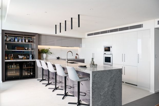 Bright, spacious contemporary kitchen with a large grey stone island, integrated white cabinetry, built-in ovens, a long breakfast bar with bar stools, and a dark built-in drinks cabinet with glass-front storage. Minimal black linear pendant lights and concealed ventilation add a clean modern finish.